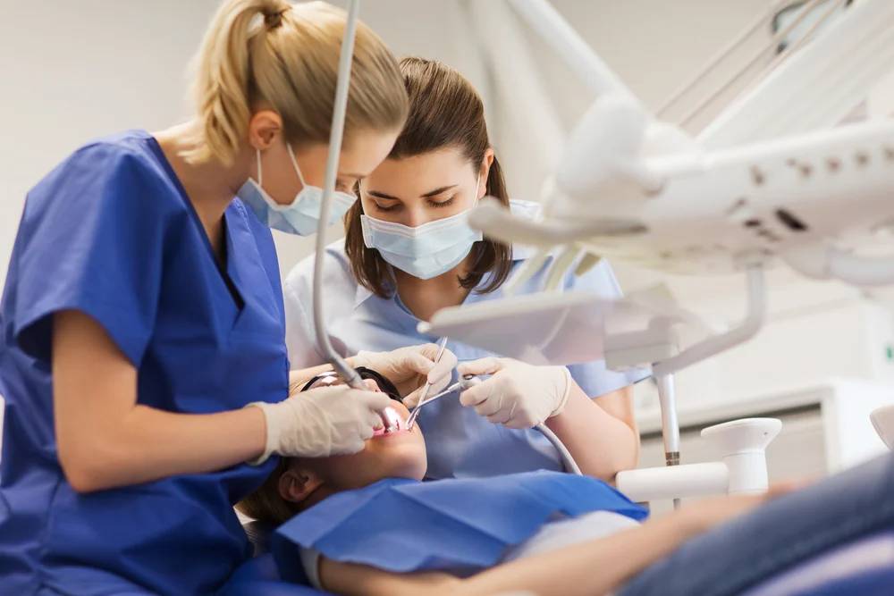 Dental professionals in blue scrubs and masks perform a procedure on a patient in a bright clinical setting